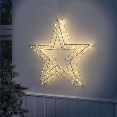 Veli Line metal lys stjerne med 960 varmt hvide LED, monteret på en hvid væg udenfor med en julegren til venstre.