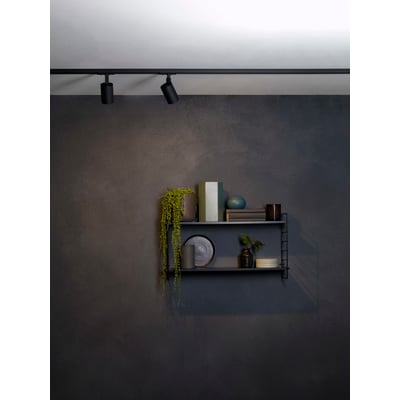 To Astro Ascoli track skinnespots i mat sort aluminium monteret på en sort skinne over en moderne hylde med dekorationer. Spottene giver stemningsfuld belysning.