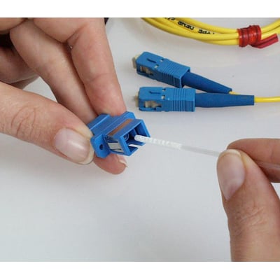 En hånd bruger en 2,5 mm fiberoptisk rensesvab med skum til at rengøre et blåt SC-fiberstik, mens andre stik ligger klar.
