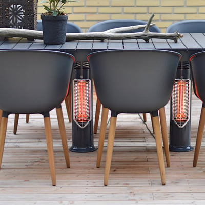 Mensa Heating IMUS terrassevarmere placeret under et havebord med stole, viser effektiv opvarmning fra lav højde på en træterrasse.