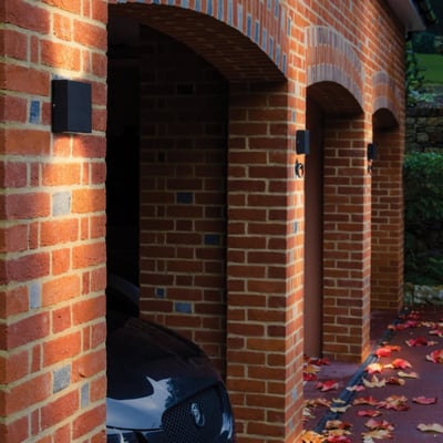 Astro 7202 Elis Twin sort væglampe monteret på en murstensvæg ved en garageindkørsel med efterårsblade på jorden.