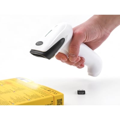 Delock 1D/2D barcode scanner i hånden, klar til at scanne en gul produktæske og viser en lille USB dongle på bordet.