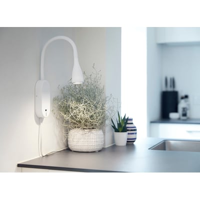 Nielsen Light Eye væglampe i hvid metal med fleksibel hals og USB-port, monteret ved siden af planter og en vase.