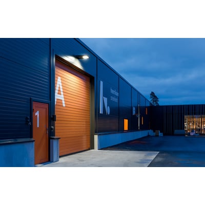 SG Armaturen Langfoss Mini projektør oplyser en stor orange garageport med bogstavet A og en mindre orange dør med tallet 1. Bygningen er mørk med blålig aftenhimmel.