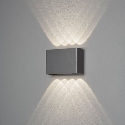 Konstsmide Chieri væglampe i aluminium, monteret på en grå væg, som sender lys opad og nedad.