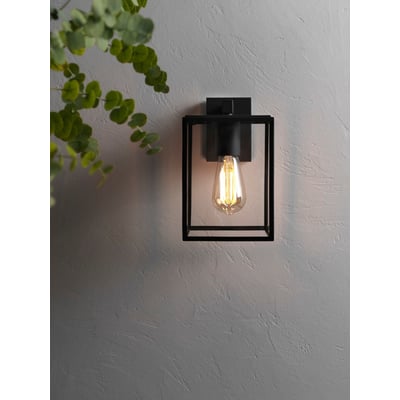Astro 8048 Box Lantern væglampe i sort sat op på en grå, tekstureret væg med en grøn plante i forgrunden.