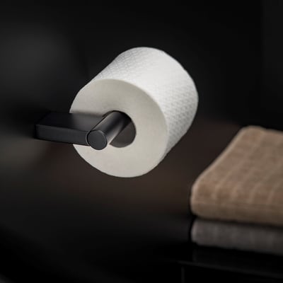 Cosmic toiletpapirholder i sort mat monteret på en væg med et toiletpapirrulle påsat. En håndklæde ses til højre.