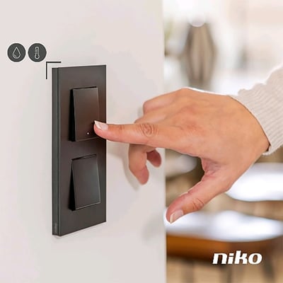 Niko 2-tryk i mørkebrun med LED og integrerede sensorer til NHC, vist i brug med en hånd der trykker.