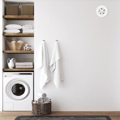 PAX Brisa hvid ventilator monteret højt på væggen over en vaskemaskine og hylder med vasketøj.
