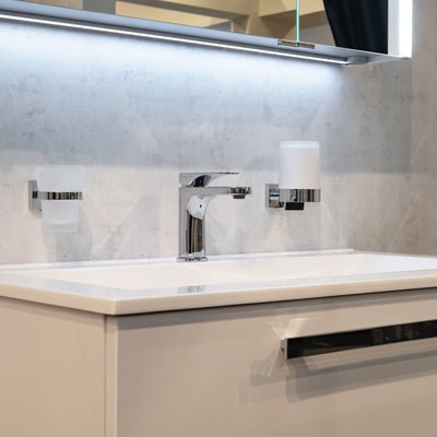 Smedbo House sæbedispenser i poleret krom med materet glas monteret på badeværelsesvæg ved siden af en vandhane og et glas.