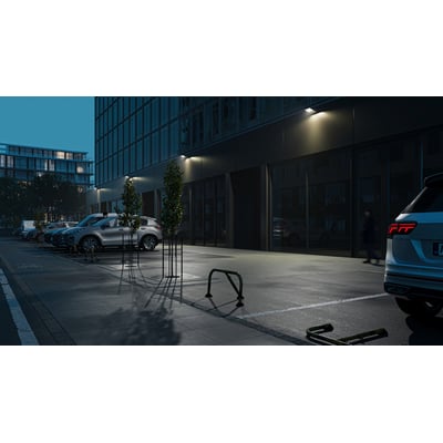 Sort Ansell Lighting WOLF væglampe med skumringsrelæ monteret på bygningsfacade om natten, oplyser en parkeringsplads.