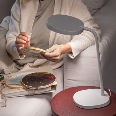 En hvid Lumie Task lysterapilampe placeret på et rundt, mørkerødt bord, der oplyser en kvinde, som sidder og broderer.