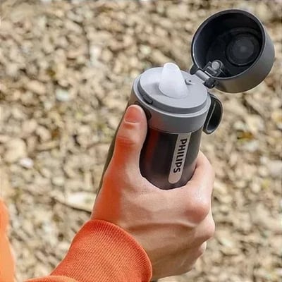 Philips filtreringstermoflaske AWP2771GRR/31 i sort, holdes af en hånd, med drikketud åben, vist udendørs i naturligt lys.