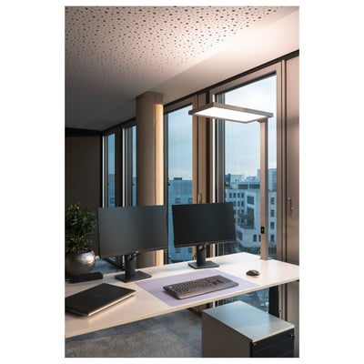 SLV Worklight TL bordlampe i sølv monteret på et skrivebord med to computerskærme. Lampen lyser neutralt hvidt og giver god arbejdsbelysning. Lampen er placeret ved siden af skærmene med udsigt til bygninger gennem vinduet.
