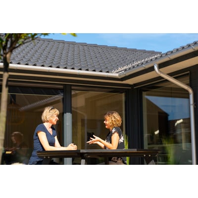 To kvinder taler sammen på en terrasse, mens Plastmo tagrenden i aluzink ses langs husets tagkant.