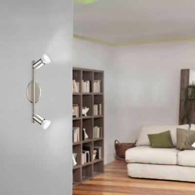 EGLO BUZZ spotlampe i børstet stål med to justerbare lamper, monteret på en grå væg i en stue med en sofa og bogreol.