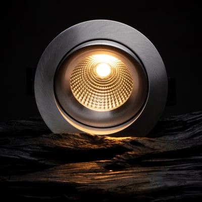 SLC One 360° downlight i aluminium med 8W LED, der lyser varmt hvidt lys op fra en reflekterende inderside på en mørk, træagtig baggrund.