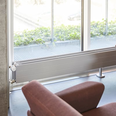 Hudevad LK lavkonvektor på 2500mm monteret langs et stort vindue med udsigt til grønne planter og et bilkørselsområde.