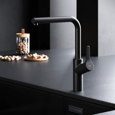AM.PM X-Joy S køkkenarmatur i mat sort messing med svingbar L-tud og etgrebsbetjening, monteret på sort bordplade med nødder i baggrunden.