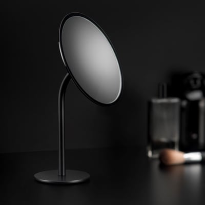Cosmic fritstående forstørrelsesspejl i sort, Ø20 cm, med elegant fod og baggrund med makeup-tilbehør.