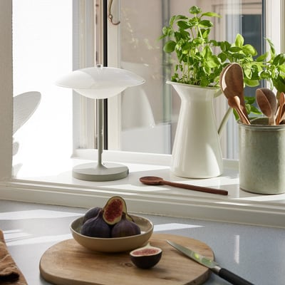 Halo Design Baroni Move bordlampe i varm grå med hvid skærm, placeret på en vindueskarm med grønne planter og frugt.