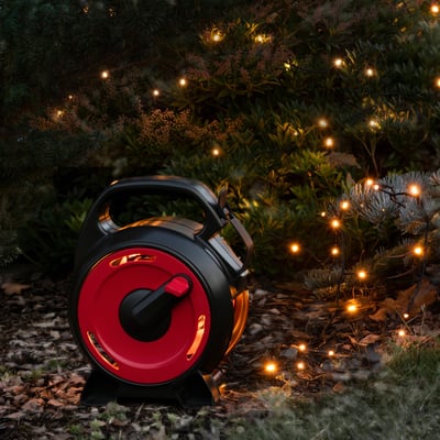 Konstsmide lyskæde med 100 varm hvide LED'er på kabeltromle, vist udendørs i en have med buske og grene.