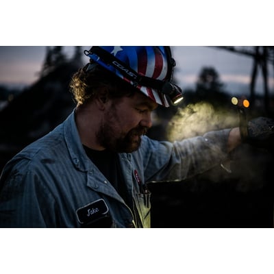 Mand med pandelampe Coast XPH34R på hovedet, oplyser arbejdsemne med kraftigt lys i mørke omgivelser.