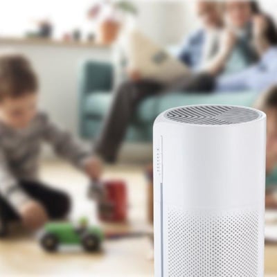 Sensibo Pure luftrenser i hvid står på et gulvtæppe, mens en familie leger og slapper af i baggrunden.