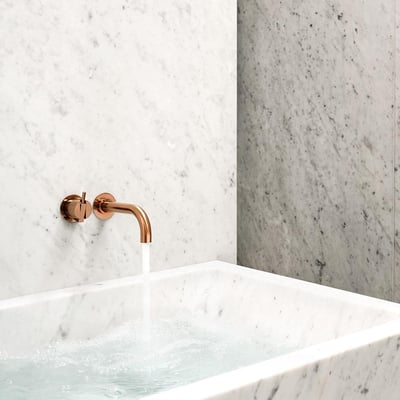 Vola 1-grebsblander SC5 i kobberfarve, monteret på marmorvæg med 360° dobbelt svingbar tud, hvor vand løber ned i et marmorbadekar.