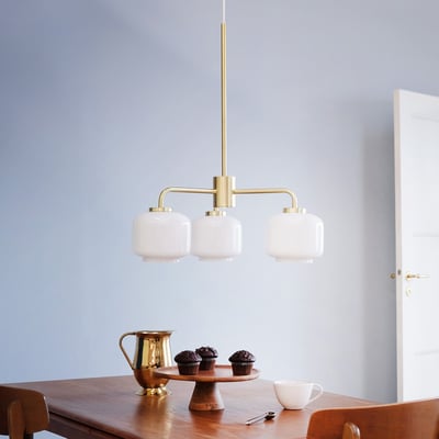 Dyberg Larsen Arp lysekrone med 3 opal glas og messing ophæng, vist over et træbord med muffins og kande.