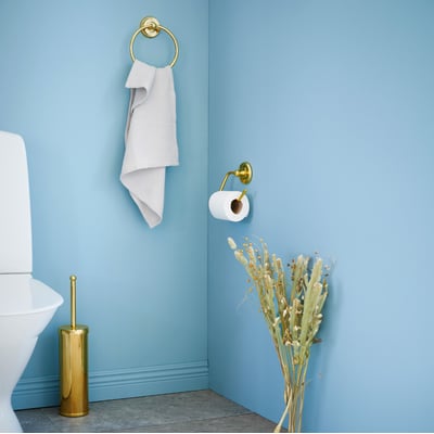 Smedbo Villa toiletpapirholder i poleret messing monteret på lyseblå væg ved siden af et toilet og et håndklædestativ.