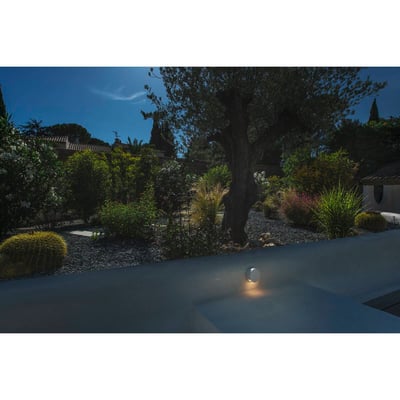 SLV Pema væglampe monteret på en facade med op/ned lysudsendelse, der oplyser en have med oliventræ og planter.