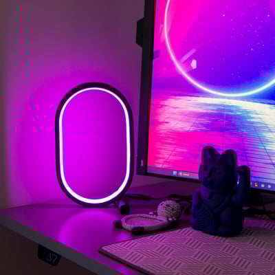 RGBIC ellipseformet lampe, 25 x 15 cm, oplyst med lilla lys på et skrivebord ved siden af en computerskærm.
