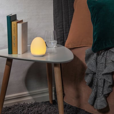 Star Trading hvid ægformet LED vågelampe, der lyser med varmt hvidt lys på et lille bord ved siden af bøger.