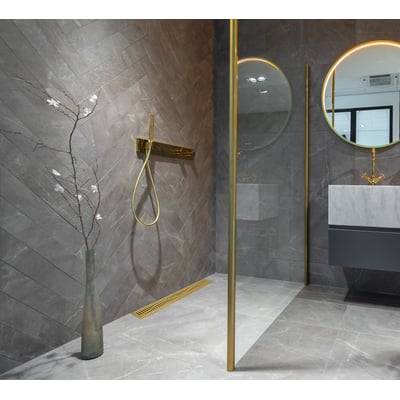 ACO ShowerDrain med Wave rist i mat guld, 105x795 mm, vist i et moderne badeværelsesmiljø med grå klinker og messing detaljer.