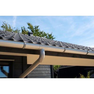 Plastmo aluzink ekspansionstudstykke til 75 mm tagrende monteret på et mørkt træbeklædt hus under en grå tagrygning og blå himmel.