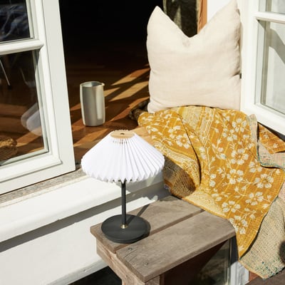 Halo Design Paris to go sort metal bordlampe med plisseret skærm placeret på et rustikt træbord med guldfarvet tæppe og pude.