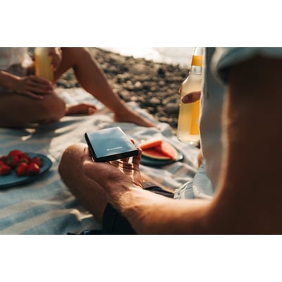 En person holder en sort Verbatim Store 'n' Go Slim Portable SSD på en strand med jordbær og drikkevarer i baggrunden.