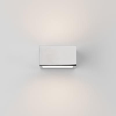 Astro 8162 Kappa LED væglampe i blank krom, tændt med varmt hvidt lys på hvid væg.