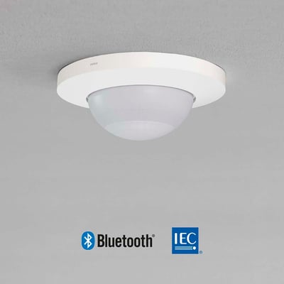 Niko P41LR tilstedeværelsessensor til indbygning i hvidt, monteret på et gråt loft med Bluetooth og IEC logoer nederst.