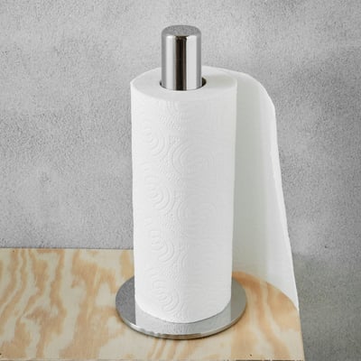 Bastant Design køkkenrulleholder i poleret rustfrit stål med en køkkenrulle monteret, placeret på en træhylde foran en grå væg.