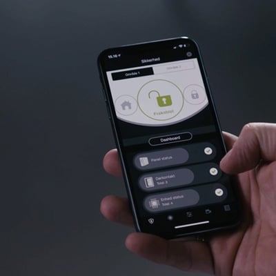 En hånd holder en iPhone med TrueGuard appen, der viser alarmens frakoblede status og status for forskellige enheder.