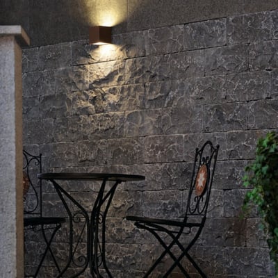 SLV Sitra væglampe i rustfarvet aluminium op/ned, monteret på en rå murstensvæg over et havebord og stole.