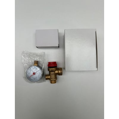 Sikkerhedsventil til 3 bar med ½" x ¾" tilslutning og trykmåler i klar plastpose, placeret ved siden af to hvide kasser.