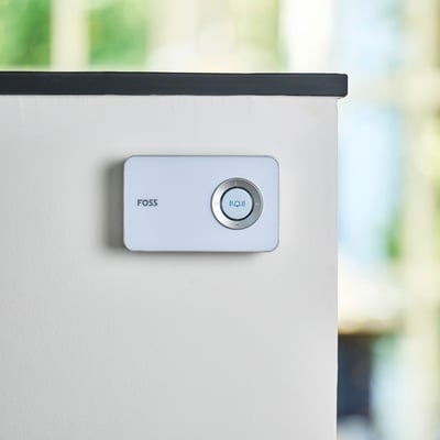 Monteret Foss Europe TREND dørklokke i hvid på en lys væg med en sort kant øverst, baggrund med grønne nuancer.