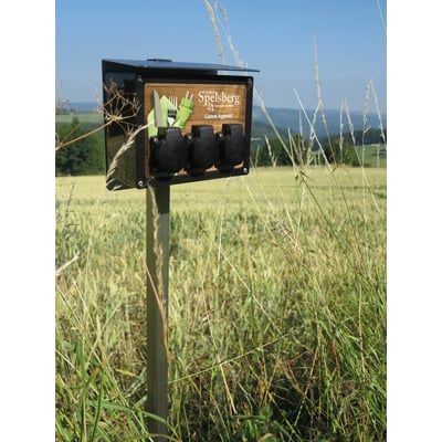 Spelsberg cykelladestation TG BCS 3 med tre sorte schuko udtag monteret på en græsmark. Stationen har logo og teksten 'Guten Appetit!'.