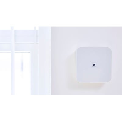 SikkertHjem S6evo™ villapakke alarmenhed monteret på en hvid væg ved siden af et vindue med hvide gardiner.