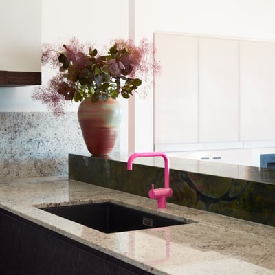 Elegant Vola KV1 køkkenarmatur i pink messing integreret i et moderne køkkenmiljø ved siden af en sort vask og et stort blomsterarrangement.