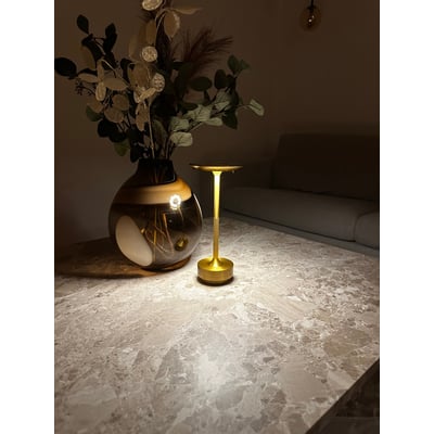 Elegant Skagen genopladelig LED lampe i guld lyser op på et marmorbord ved siden af en vase med blomster og grønne planter.