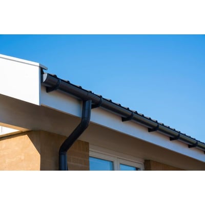 Plastmo tagrende bøjning 75 mm 60° i grafit, monteret på et hvidt facade med blå himmel i baggrunden.